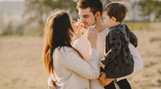 Man and woman about to kiss each other with baby in arms. Man is wearing a face mask.