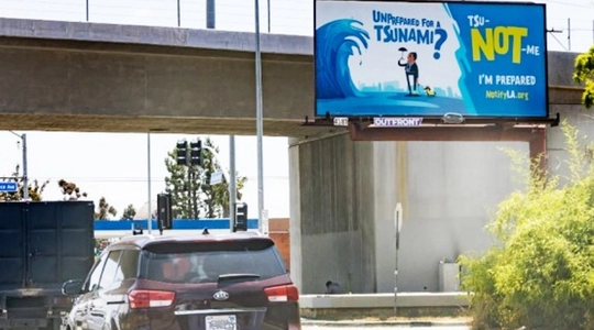 A freeway underpass is shown with traffic passing a tsunami awareness campaign billboard.