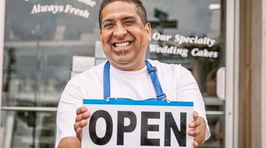 hoto of a smiling business owner wearing an apron and holding a sign that says: "Open" in front of a bakery window.