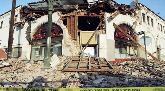 Historical photo of businesses on a corner of Hollywood Boulevard in Los Angeles with crumbled facades and a street below filled with rubble.