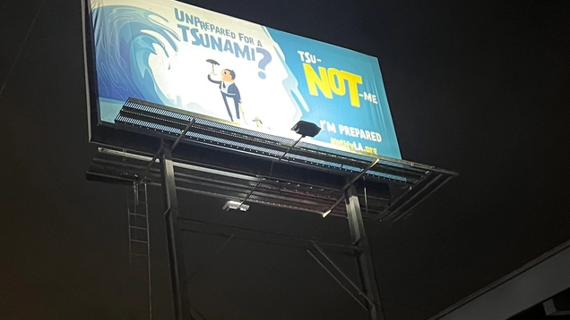Photo of a large billboard for the tsunami awareness campaign launched in May 2023, with the drawing of a cartoon figure standing under a massive ocean wave.  The caption reads "Unprepared for a Tsunami? TSu-NOT-me. NotifyLA,org" (in English and a similar play-on-words in Spanish).