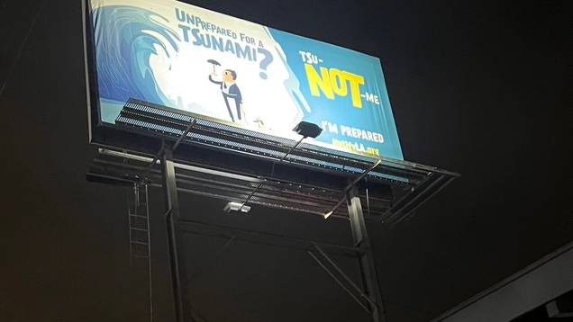 Photo of a large billboard for the tsunami awareness campaign launched in May 2023, with the drawing of a cartoon figure standing under a massive ocean wave. The caption reads "Unprepared for a Tsunami? TSu-NOT-me. NotifyLA,org" (in English and a similar play-on-words in Spanish).