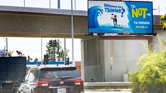 A freeway underpass is shown with traffic passing a tsunami awareness campaign billboard.