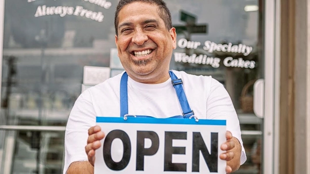 hoto of a smiling business owner wearing an apron and holding a sign that says: "Open" in front of a bakery window.