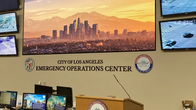 A side view of the main wall of the LA City Emergency Management Department's "second home" -- the Emergency Operations Center, in diowntown Los Angeles. A very large monitor mounted on the wall is surrounded by six smaller screens showing traffic views of key City locations. Desks and computers can be seen at the bottom and a podium for holding meetings. The logos of the City and EMD are on either side of the words on the wall EMERGENCY OPERATIONS CENTER.