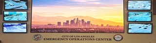 A widescreen view of the main wall of the LA City Emergency Management Department's "second home" -- the Emergency Operations Center, in downtown Los Angeles. A very large monitor mounted on the wall is surrounded by six smaller screens showing traffic views or key City locations. Desks and computers can be seen at the bottom and a podium for holding meetings. The logos of the City and EMD are on either side of the words on the wall EMERGENCY OPERATIONS CENTER.